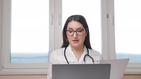 Female Doctor with Glasses and Stethoscope Consults the Patient at Online with Laptop