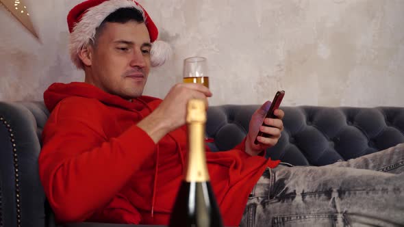 Young Man in Santa Claus Hat Browses Smartphone and Drinks Champagne Lying on Couch