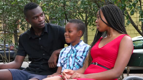 A Young Black Family Sits on a Bench in an Urban Area and Talks To Each Other