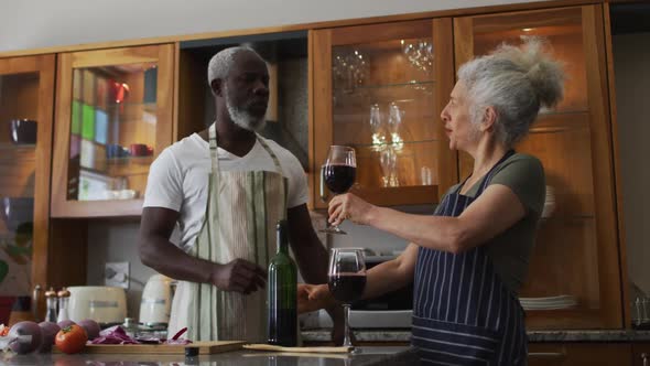 Caucasian senior woman wearing apron giving wine glass to her husband in the kitchen at home