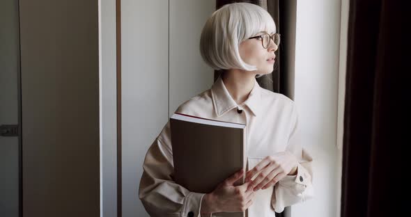 Beautiful Woman with Glasses Holds a Book in Her Hand and Looks Out the Window