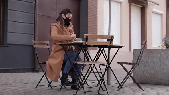 Business Woman in Face Mask Sitting Outdoor in Cafe on Terrace and Working on Laptop