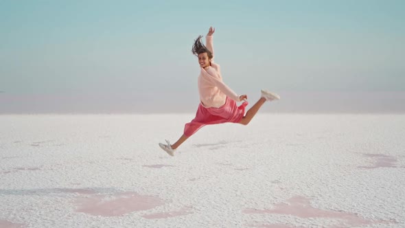 Happy Joyful Smiling Woman in Pink Oversize Wear Making Funny Jump with Raised Legs on White Salty