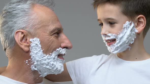 Old Man and Grandson With Shaving Foam on Faces Showing Razors Into Camera