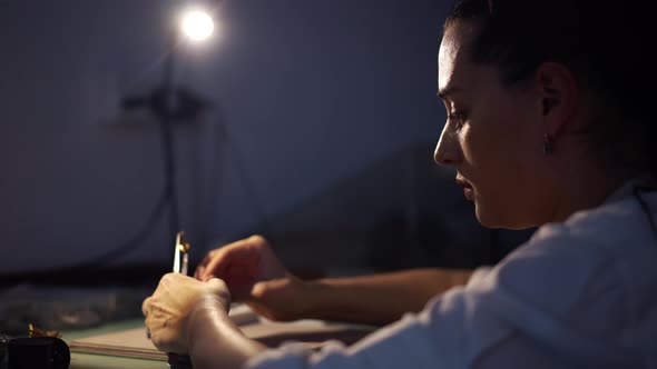 Young Female Jeweller Sitting in Workshop with Blue Light with Fire Opal in Tweezers