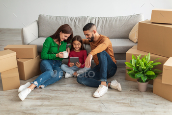 arch for household goods and furniture for their new home online on moving day. Happy young homeowners sitting on floor among cardboard boxes, plants