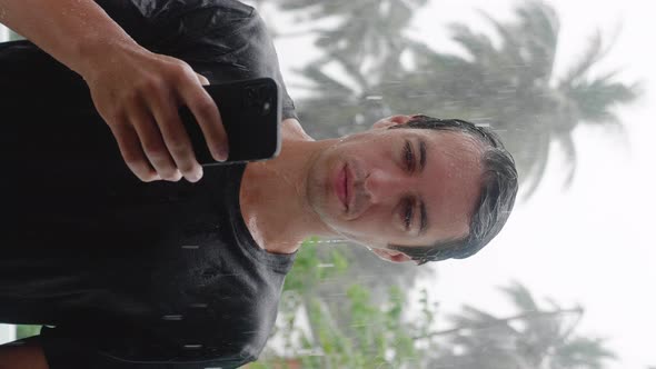 Vetrical Shot of Wet Young Man Standing in the Pouring Tropical Rain Using His Smartphone