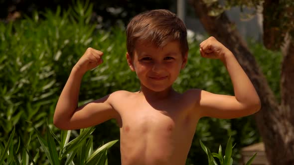Little Cheerful Boy is Smiling Happily and Showing His Biceps Muscle Outdoors