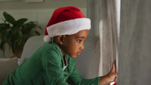 Animation of sad african american boy wearing santa hat looking outside window at christmas time