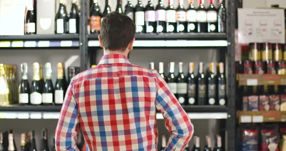 Back View of Man Scratching Head As Looking at Shelves with Bottles of Wine. Confused