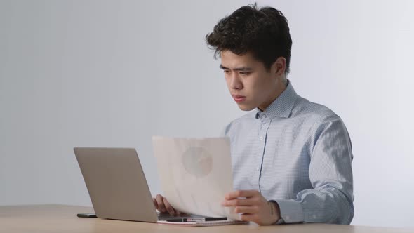 Young Concentrated Asian Businessman Reading Financial Documents with Project Chart and Thinking