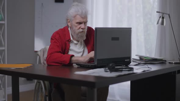 Serious Concentrated Disabled Man in Wheelchair Typing on Laptop Keyboard Messaging Online From Home