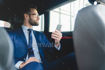man in formal clothes using smart phone while sitting at backseat of taxi car with a driver looking at the window.