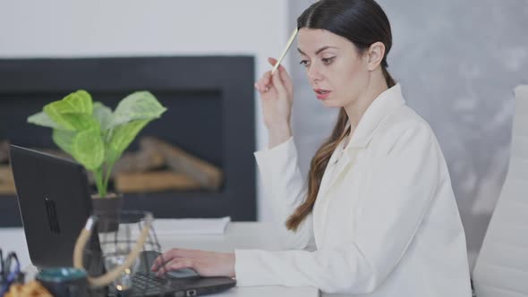 Side View of Thoughtful Young Slim Woman Surfing Internet and Messaging Online on Laptop