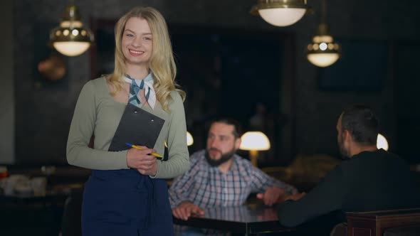 Happy Smiling Young Waitress Posing in Restaurant with Blurred Men Talking Sitting Table at