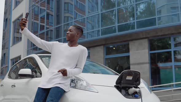 Stylish African American Man Stands Near an Electric Car Charging and Takes a Selfie with His