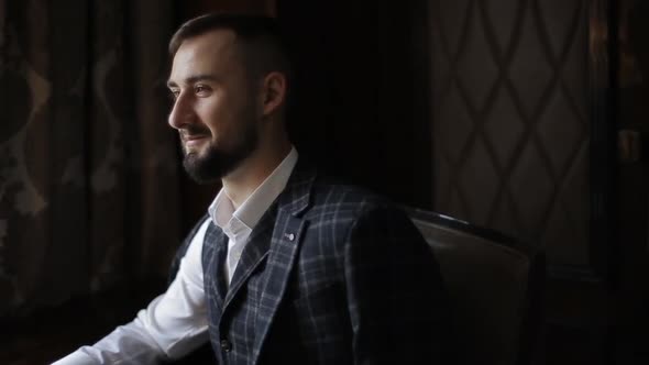 Close-up Side View of Handsome Bearded Groom in a Stylish Suit Sitting on Chair in the Morning in a