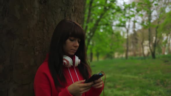 Woman stands near a tree in a park, uses a smartphone and listens to music in an earpiece.