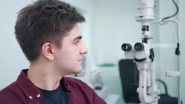 Young Confident Patient Sitting at Refractometer Looking Back at Equipment Turning Smiling