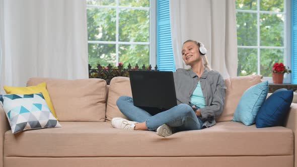 Positive Woman In Wireless Headphones Using Laptop Sitting On Couch At Home