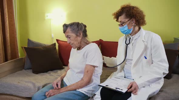 Mature Woman Doctor Holding Stethoscope Examining Senior Grandma Patient at Home