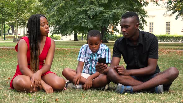 A Black Family Sits on Grass in a Park, Looks at a Smartphone and Talks About Something