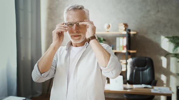 Senior Male is Smiling and Putting on His Glasses While Posing Against Blurred Background of Home