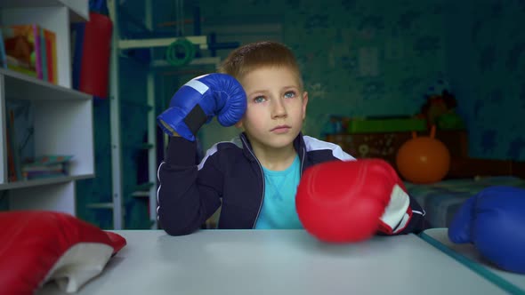 The boy dreams of playing box with friends. Sad boy in boxing gloves is sitting at the table