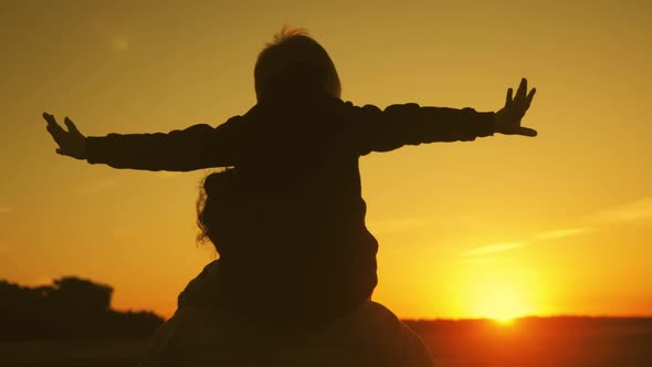 Mother and Little Son Silhouettes Playing on Meadow. Boy Raising Up Hands Imitating a Flight at