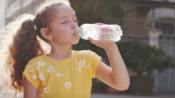 Cute Girl Child Drinks From a Plastic Bottle on the Street Near the House. Slow Motion Little Girl