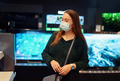 Young woman in medical face mask pensively examines variety of tv sets on display in tech store - PhotoDune Item for Sale