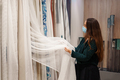 Young woman in face mask choosing tulle and curtains in fabric store, doing shopping during covid19 - PhotoDune Item for Sale