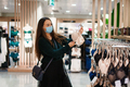 Young beautiful woman wearing face mask standing in lingerie store department, choosing bra - PhotoDune Item for Sale