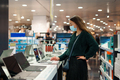 Focused young woman in face mask examines laptop on display in tech store department - PhotoDune Item for Sale