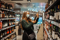 Young cheerful woman in medical face mask shopping during pandemic standing in wine store - PhotoDune Item for Sale