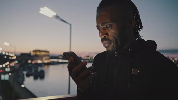 Focused African American Man Reading Message on Smartphone Standing on Bridge in Evening Tracking