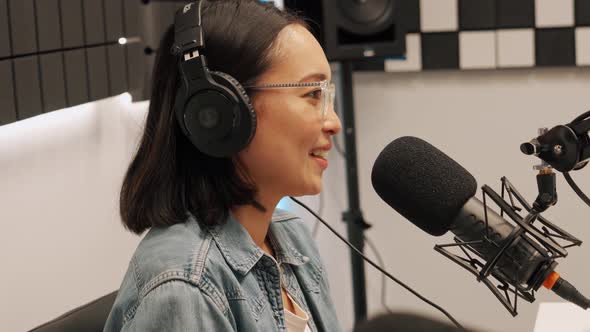 Laughing young Asian woman talking into microphone while recording radio