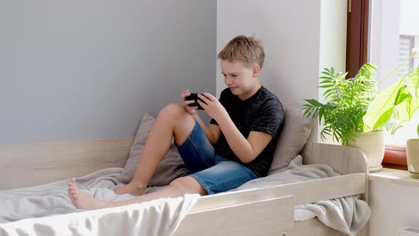 Boy Playing Mobile Game on Smartphone Sitting on Bed in His Room