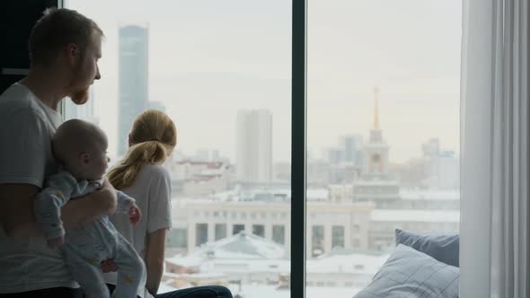 family with toddler kiss each other standing by the window overlooking the city