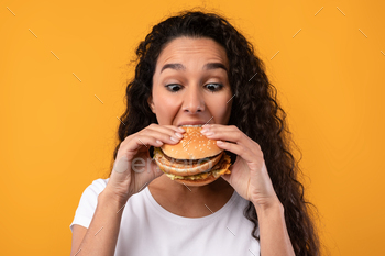 r Eating Junk Food Posing Over Yellow Orange Studio Background. Woman Enjoying Big Hamburger. Unhealthy Nutrition And Binge Eating Habit Concept