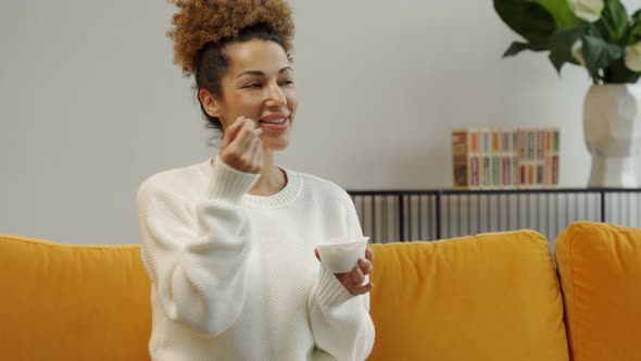 Beautiful Black Woman Eating Yogurt As Healthy Breakfast or Snack at Home Sitting on the Couch