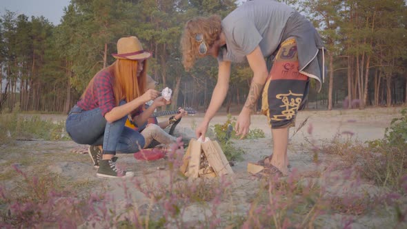 Young Hipster Guy and His Girlfriend Making a Fire with Woods on the Background of Their Friend