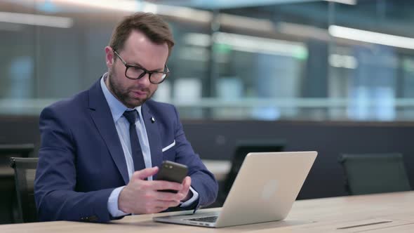 Middle Aged Businessman with Laptop Using Smartphone