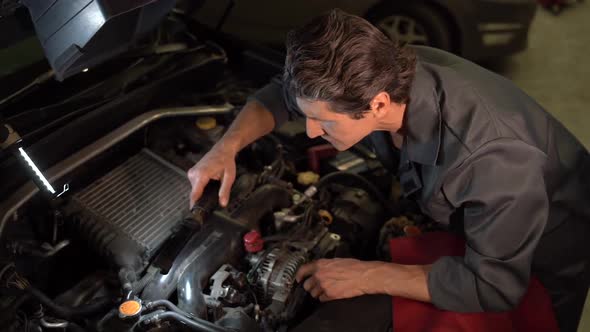 High angle view of the auto mechanic standing at the opened hood and lighting with lamp