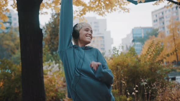 Happy Young Blond Woman with Hoodie Listening To the Music on Heaset and Dancing in the City Park in