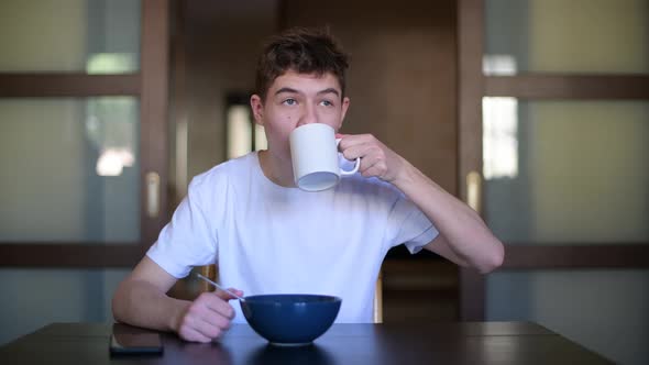 A teenager eats porridge and drinks tea, looking out the window
