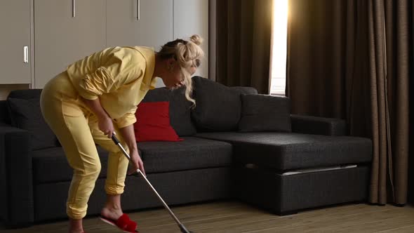 Happy Blonde Woman in a Yellow Hoodie with a Mop Cleaning the House and Dancing