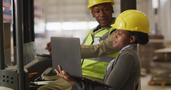 African american male and female workers wearing safety suits and using laptop in warehouse