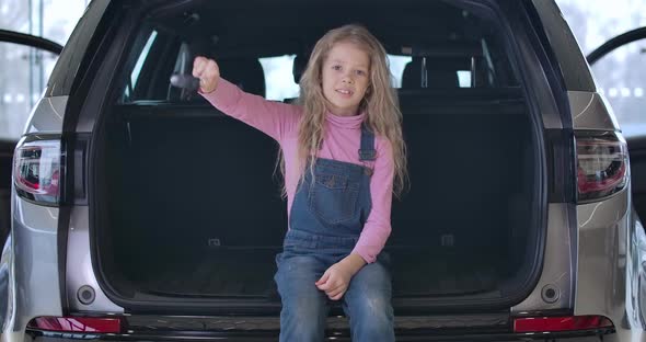 Portrait of Cheerful Caucasian Little Girl Sitting in Car Trunk and Bragging Keys. Charming Blond