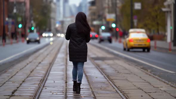 a Brunette with Long Hair in a Knitted Sweater and a Dark Coat Walks Along the Tram Tracks on a